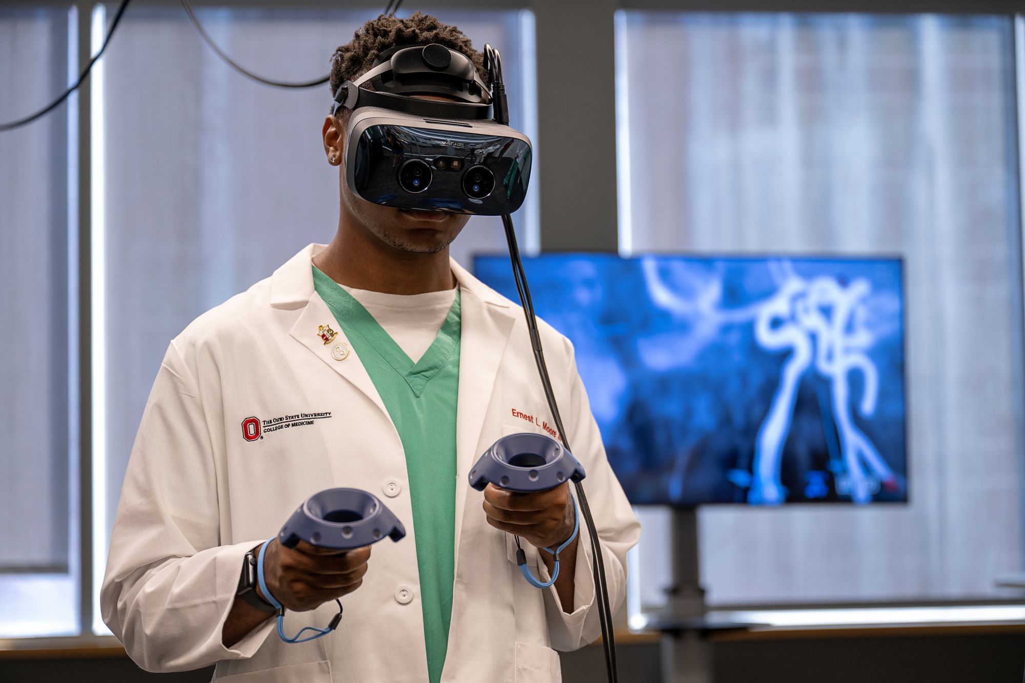 Male student in a lab coat, wearing VR headset and holding controllers, engaging with anatomy that is on a TV behind him.