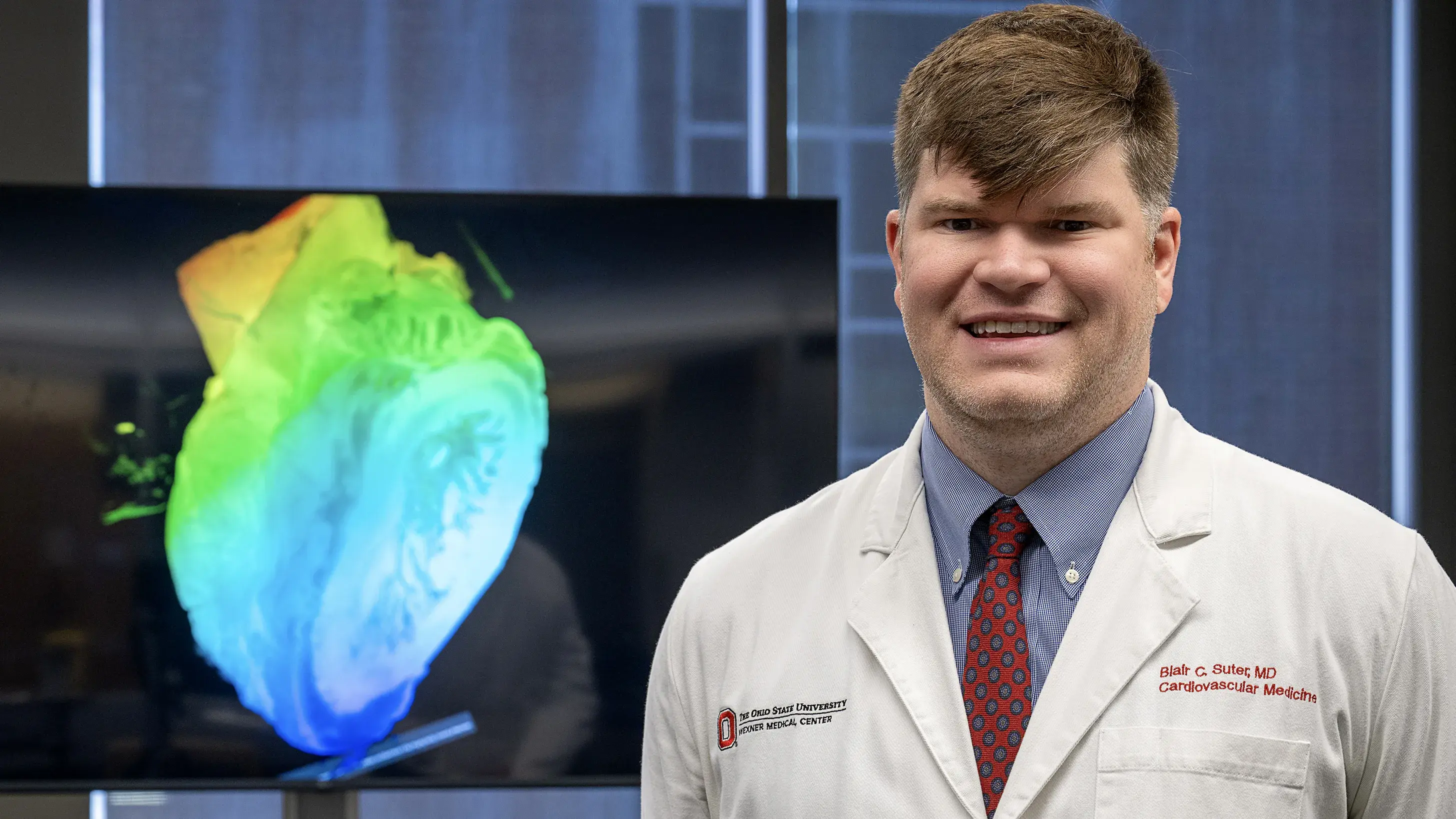 Dr. Blair Suter standing and smiling in front of a anatomical heart on a display.
