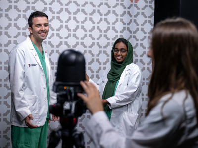 Three medical professionals in white coats pose in front of a patterned wall, smiling. A camera is set up, capturing the scene, creating a friendly and professional atmosphere.