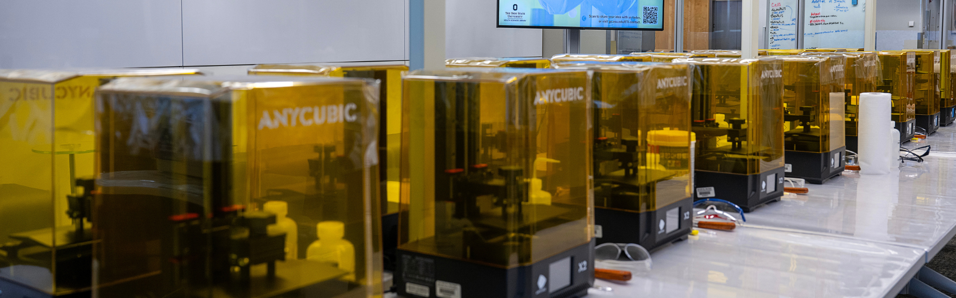 Two rows of 3D printers with covers are lined up on a table.