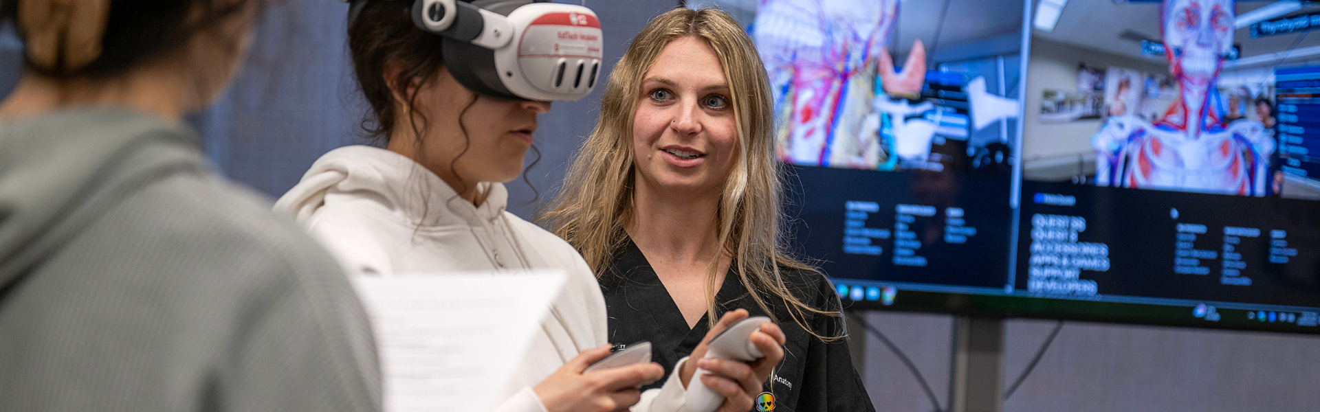 An Ohio State faculty member assists a student wearing a VR headset and hand controllers during a classroom exercise