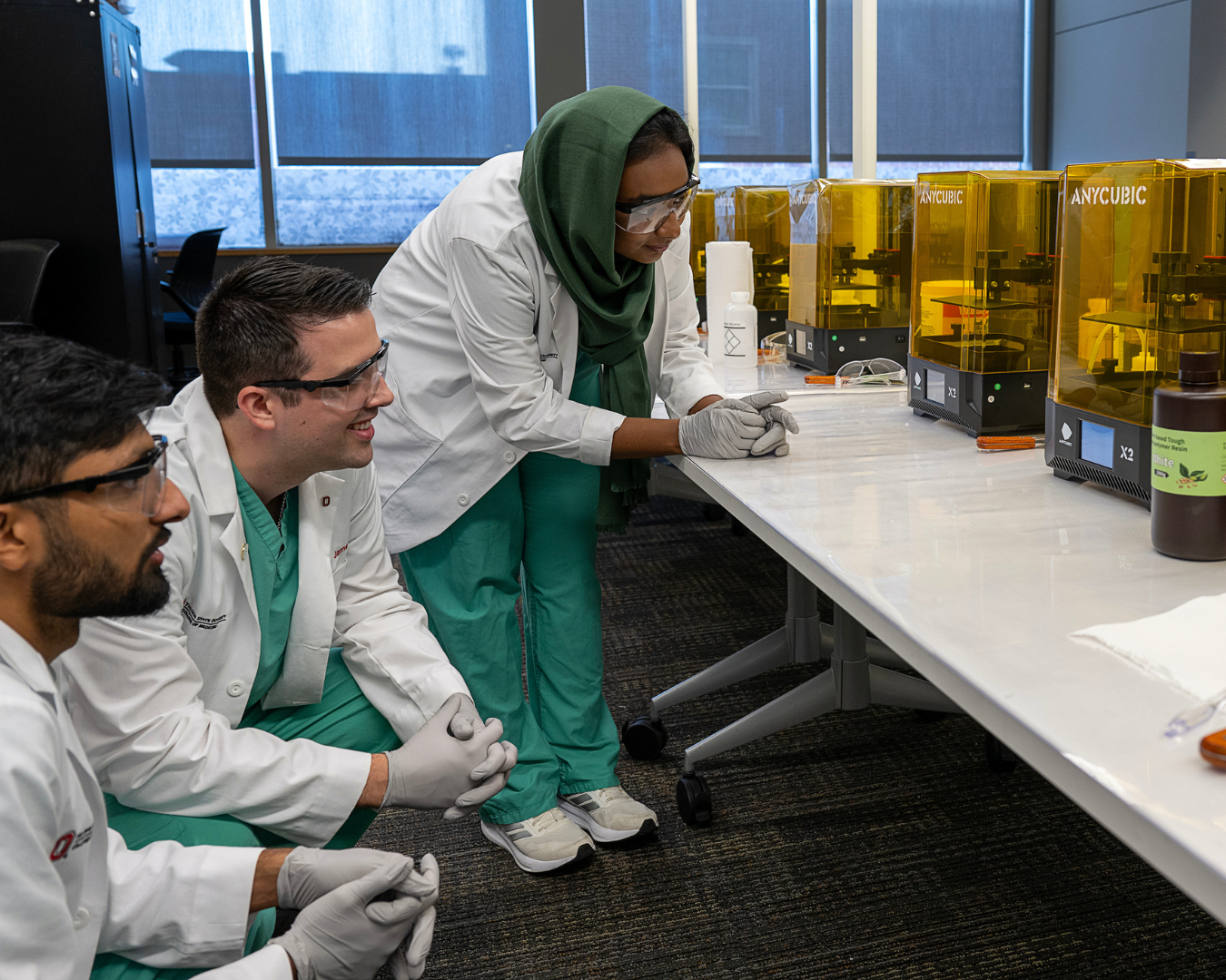 Three Ohio State students in lab coats and safety equipment observing the progress on a 3D printer.